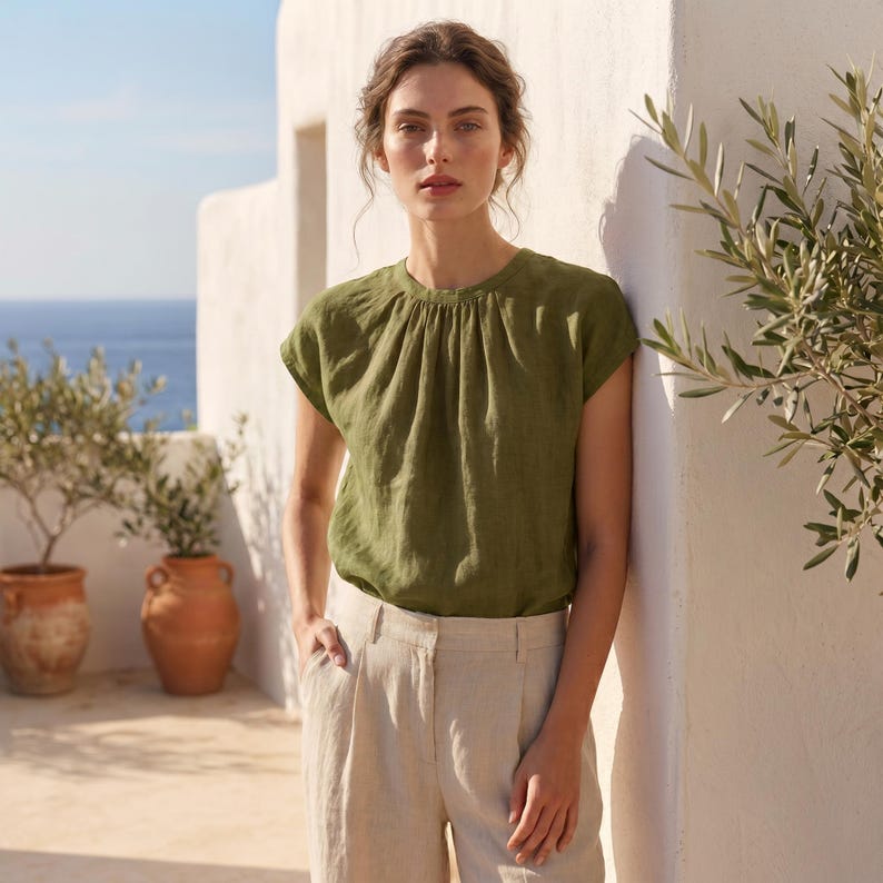 Peut inclure: Haut en lin vert olive avec encolure fronc&eacute;e et manches courtes. Port&eacute; avec un pantalon en lin beige clair. Le mod&egrave;le se tient &agrave; l'ext&eacute;rieur pr&egrave;s d'un mur blanc et de pots en terre cuite avec des oliviers. L'arri&egrave;re-plan montre la mer et le ciel bleu.