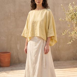 May include: A woman wearing a light yellow top with wide sleeves and a cream-colored wrap skirt. The outfit is made of a lightweight fabric. The background features a neutral-toned wall and a small tree.