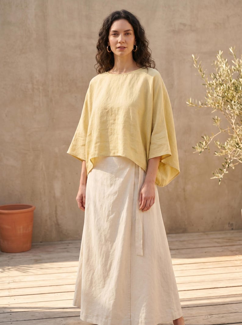 May include: A woman wearing a light yellow top with wide sleeves and a cream-colored wrap skirt. The outfit is made of a lightweight fabric. The background features a neutral-toned wall and a small tree.