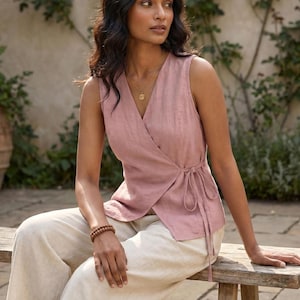 A woman in a pink sleeveless top and beige pants sits on a wooden bench, looking off to the side with a thoughtful expression.