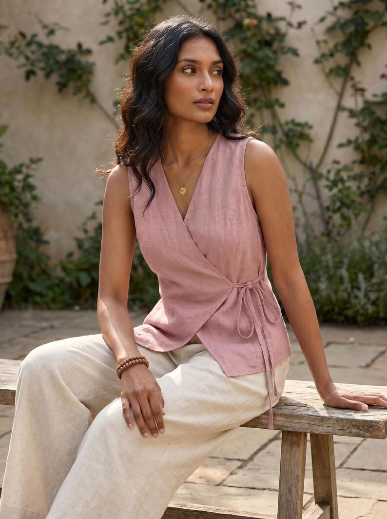 A woman in a pink sleeveless top and beige pants sits on a wooden bench, looking off to the side with a thoughtful expression.