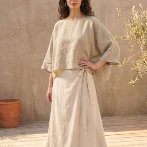 May include: A beige linen outfit consisting of a top and a wrap skirt. The top has a loose, flowing design with wide sleeves. The skirt wraps around the waist and ties with a matching fabric belt. The outfit is worn outdoors.