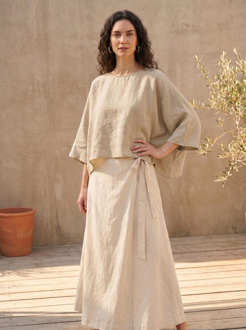 May include: A beige linen outfit consisting of a top and a wrap skirt. The top has a loose, flowing design with wide sleeves. The skirt wraps around the waist and ties with a matching fabric belt. The outfit is worn outdoors.
