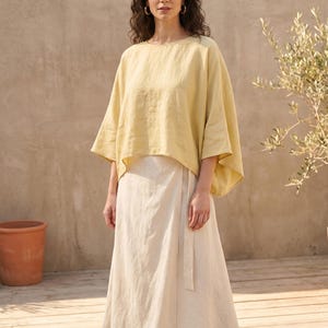 May include: A woman wearing a yellow linen top and a cream-colored wrap skirt. The top has wide sleeves and a round neckline. The skirt is long and flowing, with a matching belt. The outfit is simple and elegant, perfect for warm weather.