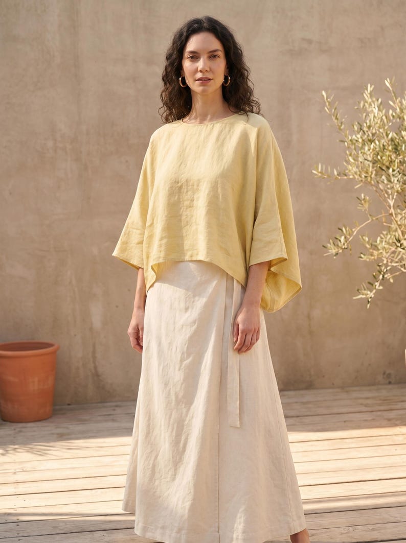 May include: A woman wearing a yellow linen top and a cream-colored wrap skirt. The top has wide sleeves and a round neckline. The skirt is long and flowing, with a matching belt. The outfit is simple and elegant, perfect for warm weather.