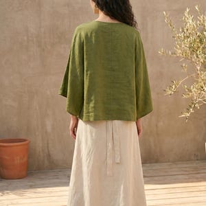 May include: A woman wearing a green linen top with wide sleeves and a long, cream-colored skirt. The outfit is simple and elegant, suitable for casual wear. The background features a wooden deck and a terracotta pot.