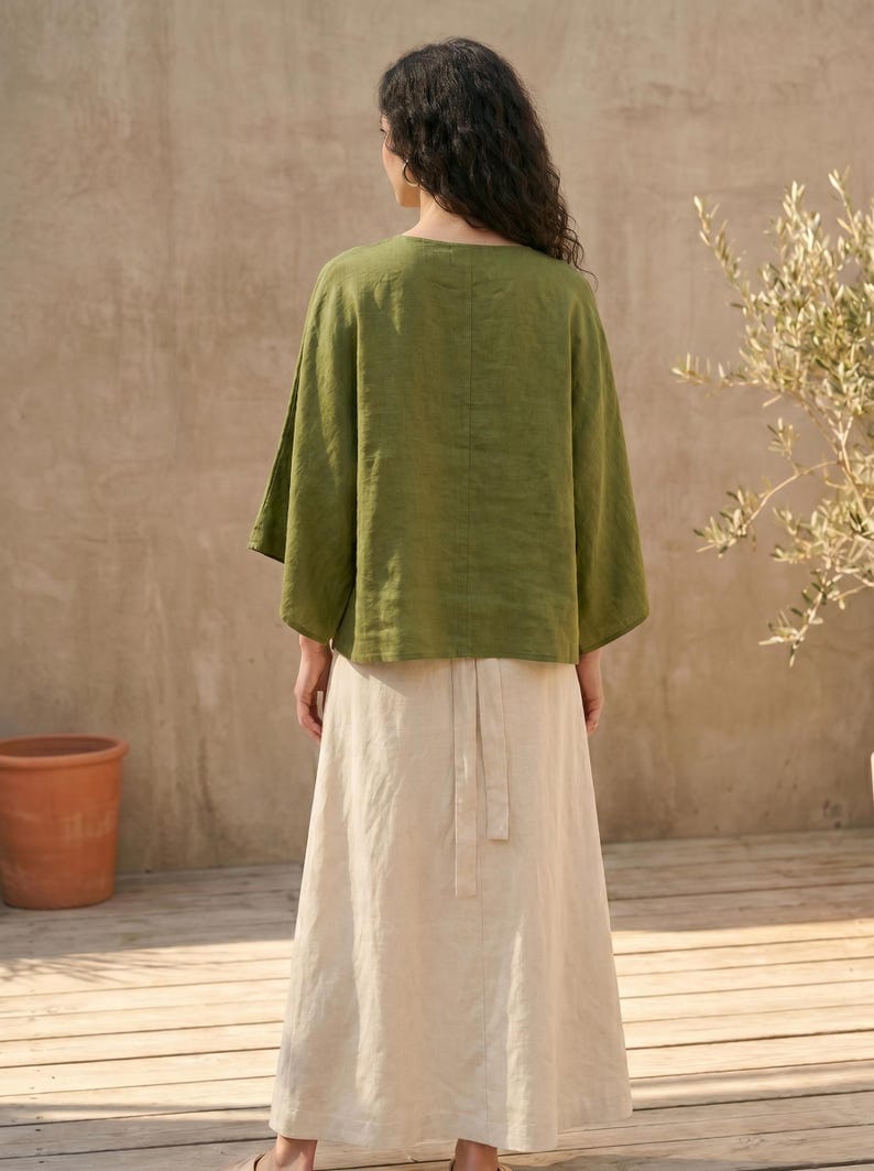 May include: A woman wearing a green linen top with wide sleeves and a long, cream-colored skirt. The outfit is simple and elegant, suitable for casual wear. The background features a wooden deck and a terracotta pot.