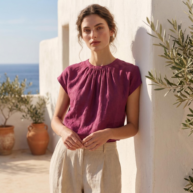 Peut inclure: Un haut en lin magenta avec un col fronc&eacute; et des manches courtes. Le haut est associ&eacute; &agrave; un pantalon en lin beige. Le mod&egrave;le est appuy&eacute; contre un mur blanc, avec la mer en arri&egrave;re-plan.