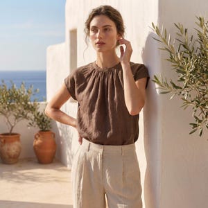 Peut inclure: Une femme porte un haut en lin marron avec un col fronc&eacute; et des manches courtes, associ&eacute; &agrave; un pantalon en lin beige clair. L'ensemble est pos&eacute; contre un mur blanc avec vue sur la mer et le ciel.