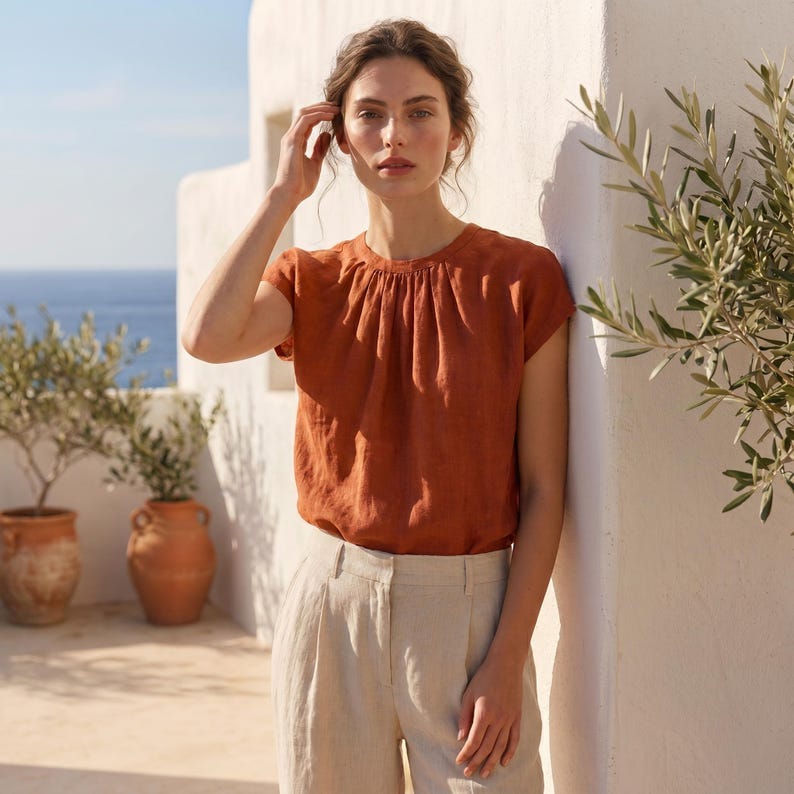 Peut inclure: Un haut &agrave; manches courtes couleur rouille avec un col fronc&eacute;. Le haut est associ&eacute; &agrave; un pantalon en lin beige clair. La sc&egrave;ne se d&eacute;roule en ext&eacute;rieur avec un mur blanc et des pots en terre cuite avec des plantes.