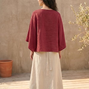 May include: A woman wearing a burgundy linen top with wide sleeves and a cream-colored linen skirt. The skirt has a high waist and a tie detail at the back. The outfit is simple and elegant, suitable for warm weather.
