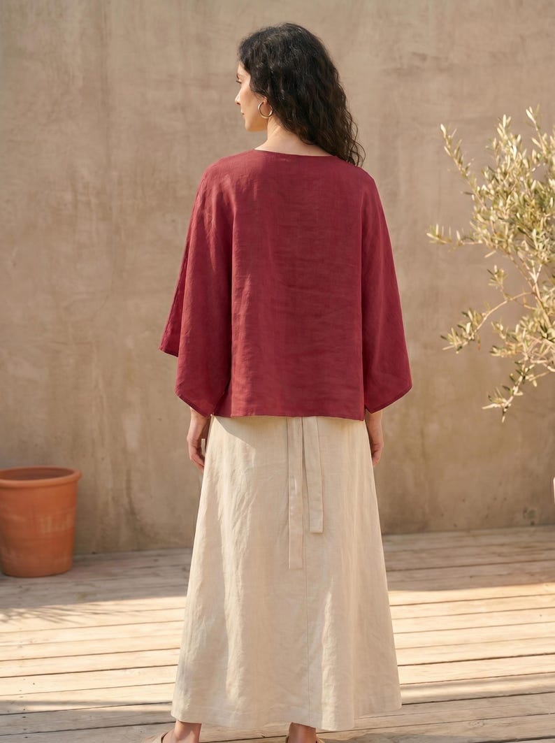 May include: A woman wearing a burgundy linen top with wide sleeves and a cream-colored linen skirt. The skirt has a high waist and a tie detail at the back. The outfit is simple and elegant, suitable for warm weather.