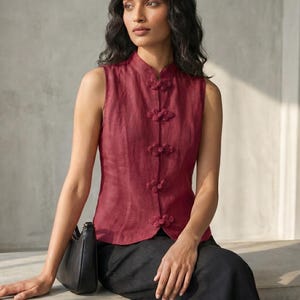 May include: A sleeveless, burgundy top with a mandarin collar and decorative frog closures. The top is paired with black pants. A black handbag rests on the model's lap.