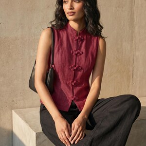 May include: A sleeveless, burgundy top with a mandarin collar and decorative frog closures. The top is paired with black, wide-leg pants. A black shoulder bag is visible. The model is sitting on a concrete block.