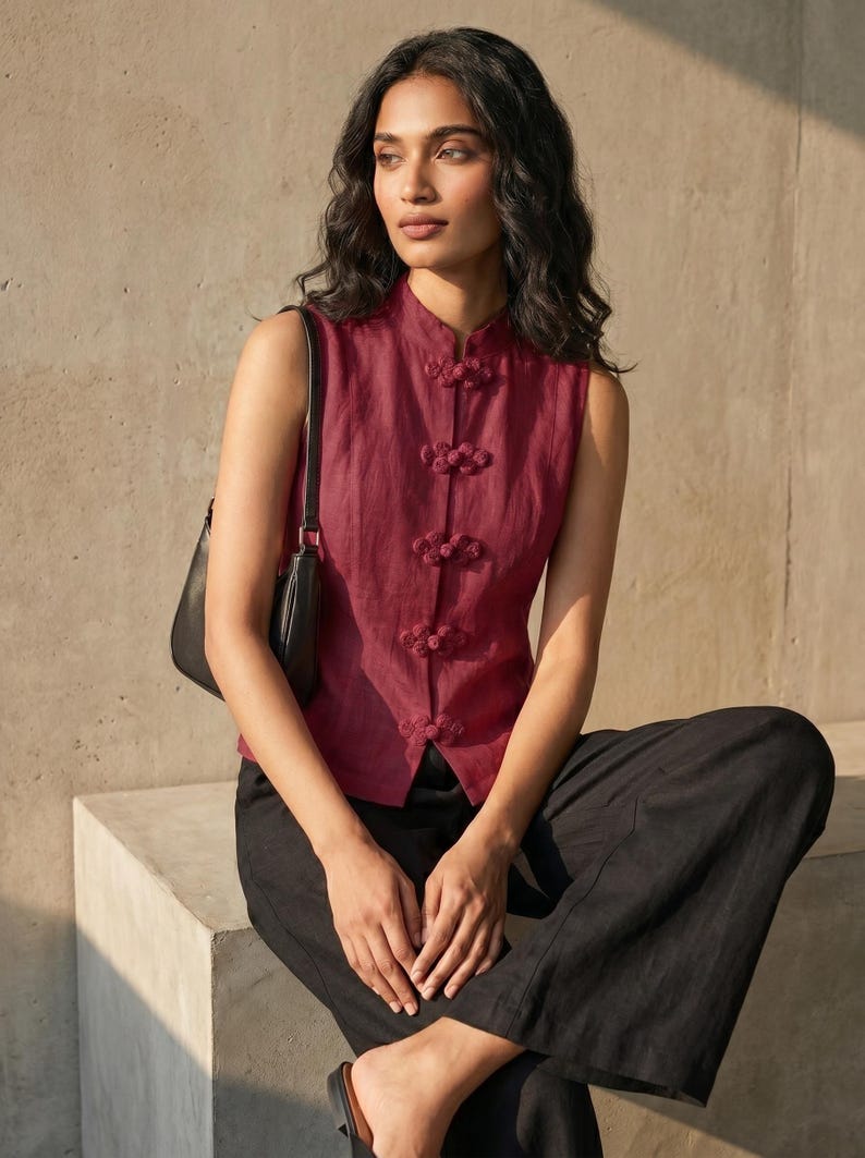 May include: A sleeveless, burgundy top with a mandarin collar and decorative frog closures. The top is paired with black, wide-leg pants. A black shoulder bag is visible. The model is sitting on a concrete block.