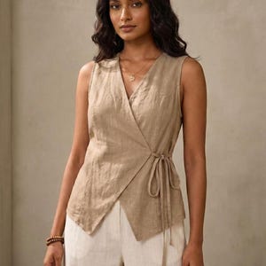 A woman with long dark hair wearing a beige sleeveless top with a wrap-style neckline and white pants.