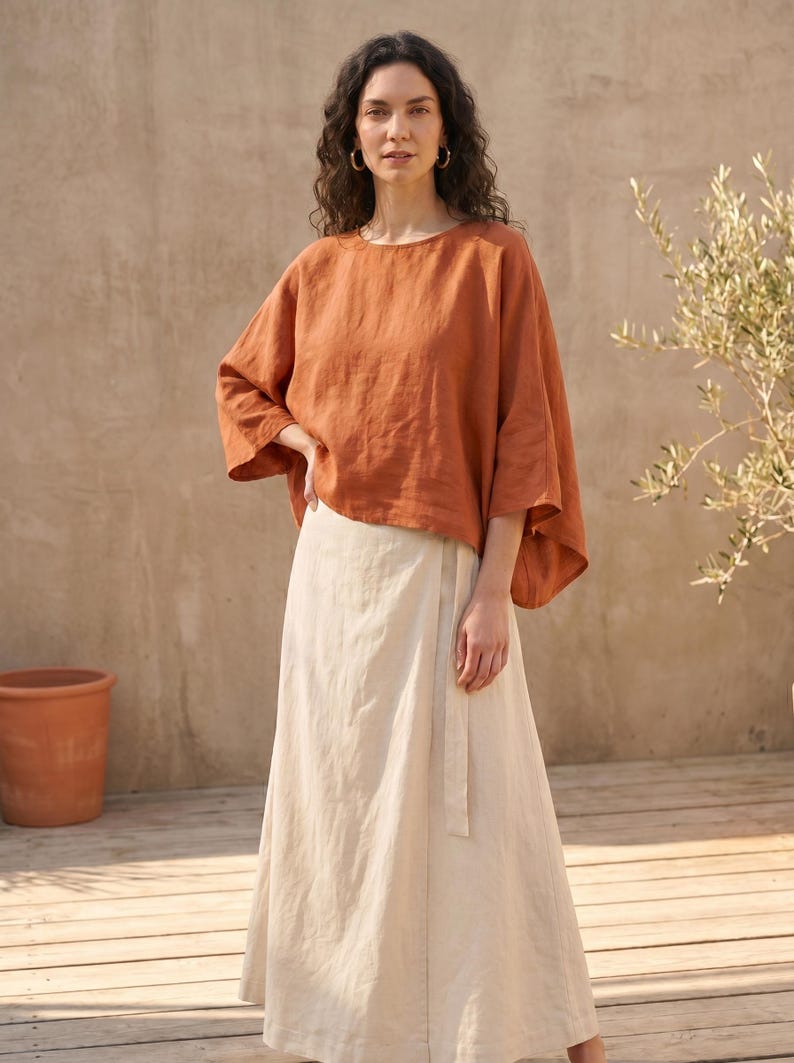 May include: A burnt orange, loose-fitting top with three-quarter sleeves and a cream-coloured wrap skirt. The outfit is made of a lightweight fabric. The model is wearing gold hoop earrings. The background includes a terracotta pot and a small tree.