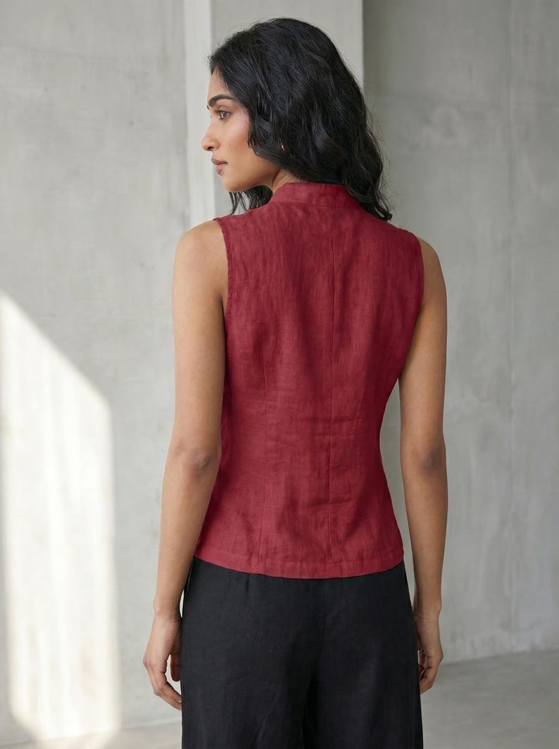 May include: A sleeveless, burgundy-colored top with a high collar. The top is made of a textured fabric and has a fitted silhouette. The model is also wearing black pants. The background is a plain, light gray wall.