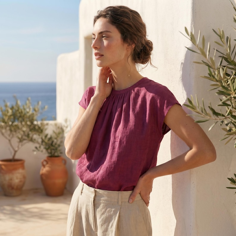 May include: A short-sleeved, magenta-colored top with a gathered neckline. The top is paired with beige-colored pants. The background features a white wall, potted plants, and a view of the ocean.