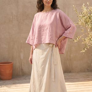 May include: A woman wearing a light pink linen top with wide sleeves and a beige wrap skirt. The outfit is made of natural fabrics and is suitable for warm weather. The background includes a wooden deck, a terracotta pot, and a plant.