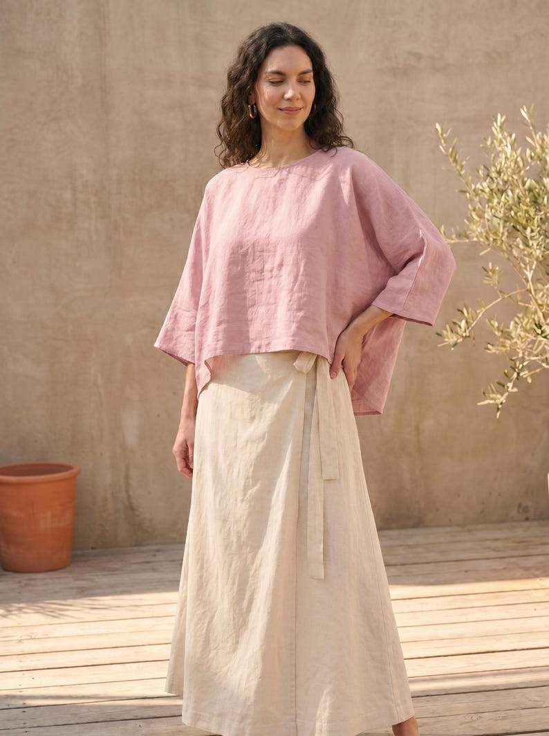 May include: A woman wearing a light pink linen top with wide sleeves and a beige wrap skirt. The outfit is made of natural fabrics and is suitable for warm weather. The background includes a wooden deck, a terracotta pot, and a plant.