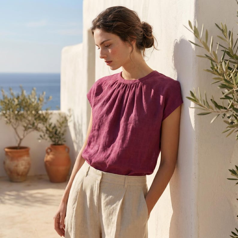 Peut inclure: Un haut magenta &agrave; manches courtes avec un col fronc&eacute;, associ&eacute; &agrave; un pantalon en lin beige clair. La sc&egrave;ne se d&eacute;roule en ext&eacute;rieur, avec un mur blanc, des plantes en pot et une vue sur la mer en arri&egrave;re-plan.