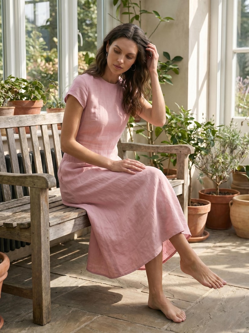 May include: A light pink linen dress with short sleeves and a round neckline. The dress is knee-length and is worn by a person sitting on a wooden bench. The background includes potted plants and a window.