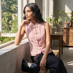 May include: A sleeveless pink top with decorative knot closures, paired with black pants and a black handbag. The top has a mandarin collar. The outfit is worn in a room with a window and wooden furniture.