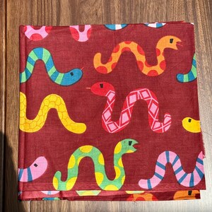 Könnte beinhalten: Ein gefaltetes Stoffquadrat mit einem sich wiederholenden Muster aus bunten Cartoon-Schlangen auf einem weinroten Hintergrund. Die Schlangen sind in verschiedenen Farben, darunter Gelb, Blau, Orange, Rosa und Grün. Der Stoff scheint Baumwolle oder ein ähnliches Material zu sein.