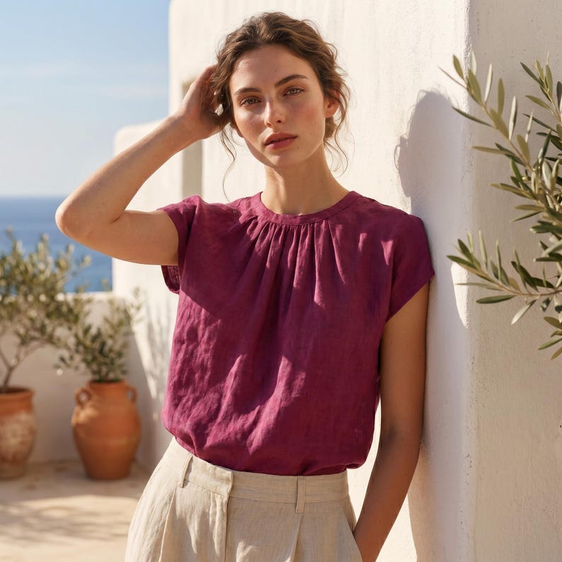 May include: A magenta-coloured short-sleeved top with a gathered neckline, worn with light beige trousers. The background includes a white wall, olive trees in terracotta pots, and a view of the sea.