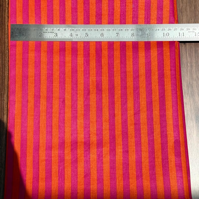 Op de afbeelding: Een levendige stof met verticale strepen in afwisselende tinten oranje en felroze. Een metalen liniaal ligt bovenop en toont de afmetingen in centimeters. Het patroon van de stof is consistent en cre&euml;ert een gedurfd visueel effect.