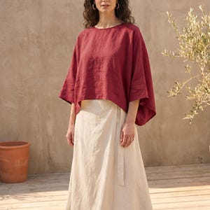 May include: A woman wearing a burgundy-colored linen top with wide sleeves and a cream-colored wrap skirt. The outfit is simple and elegant, suitable for warm weather. The background features a neutral-toned wall and wooden deck.