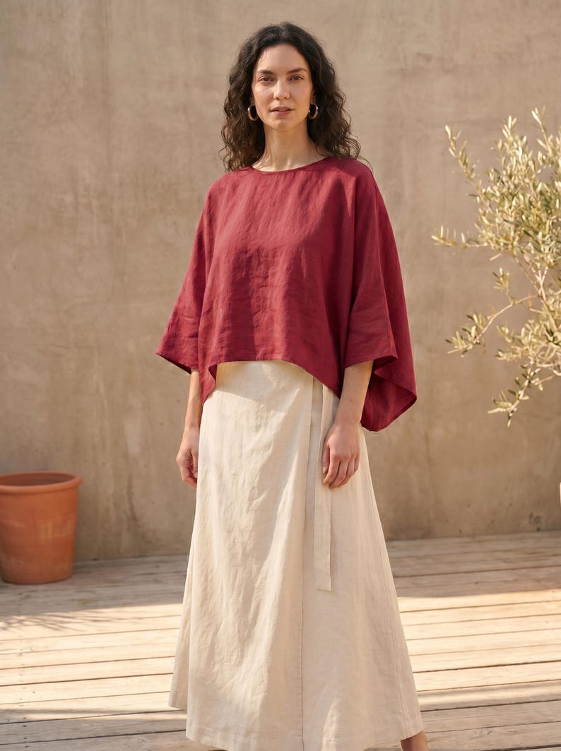May include: A woman wearing a burgundy-colored linen top with wide sleeves and a cream-colored wrap skirt. The outfit is simple and elegant, suitable for warm weather. The background features a neutral-toned wall and wooden deck.