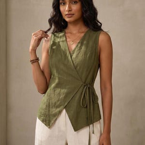 A woman with long dark hair wearing a green sleeveless top and white pants, posing against a beige background.