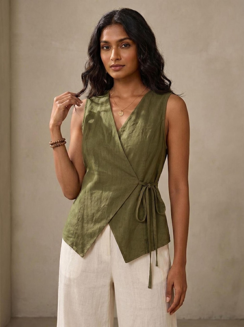 A woman with long dark hair wearing a green sleeveless top and white pants, posing against a beige background.