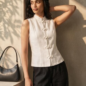 May include: A sleeveless white top with a mandarin collar and decorative knot closures. The top is paired with black pants and a black handbag. The outfit is simple and elegant, suitable for various occasions.