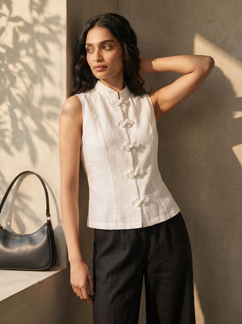 May include: A sleeveless white top with a mandarin collar and decorative knot closures. The top is paired with black pants and a black handbag. The outfit is simple and elegant, suitable for various occasions.