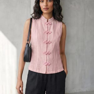 May include: A sleeveless, light pink top with a mandarin collar and decorative flower-shaped buttons. The top is paired with black pants and a black shoulder bag. The outfit is simple and elegant.