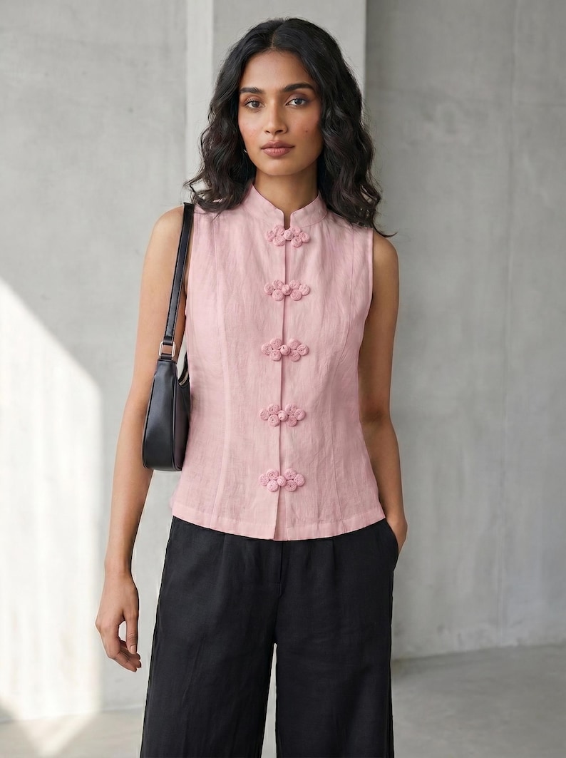May include: A sleeveless, light pink top with a mandarin collar and decorative flower-shaped buttons. The top is paired with black pants and a black shoulder bag. The outfit is simple and elegant.