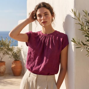 May include: A short-sleeved, magenta-colored top with a gathered neckline. The top is paired with light beige pants. The background features a white wall, olive trees, and a glimpse of the ocean.