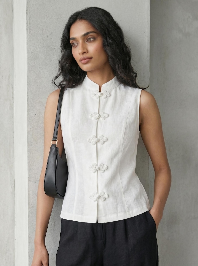 May include: White sleeveless top with a mandarin collar and decorative floral buttons. The top is paired with black pants and a black shoulder bag. The style is minimalist and elegant.