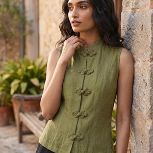 May include: Olive green sleeveless top with decorative frog closures. The top has a mandarin collar and is made of a lightweight fabric. The model is wearing black pants. The background includes a stone wall and greenery.