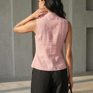 May include: A sleeveless, light pink linen top with a mandarin collar. The top is paired with black pants. The model is holding a black handbag. The outfit is simple and elegant.