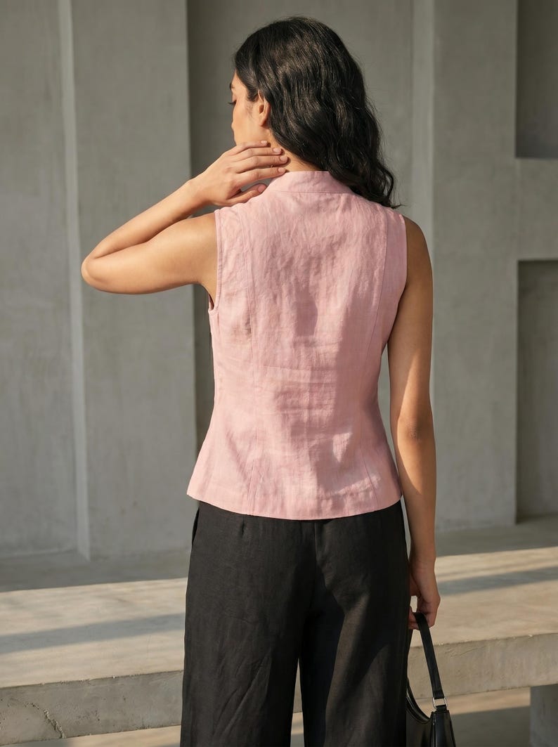 May include: A sleeveless, light pink linen top with a mandarin collar. The top is paired with black pants. The model is holding a black handbag. The outfit is simple and elegant.