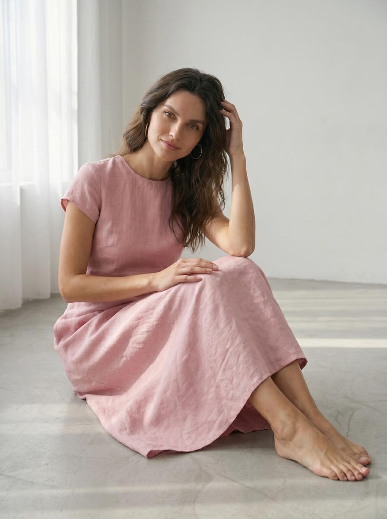 May include: A pale pink linen dress with a round neckline and short sleeves. The midi-length dress is worn by a person sitting on the floor. The dress is made of a soft, flowing fabric.