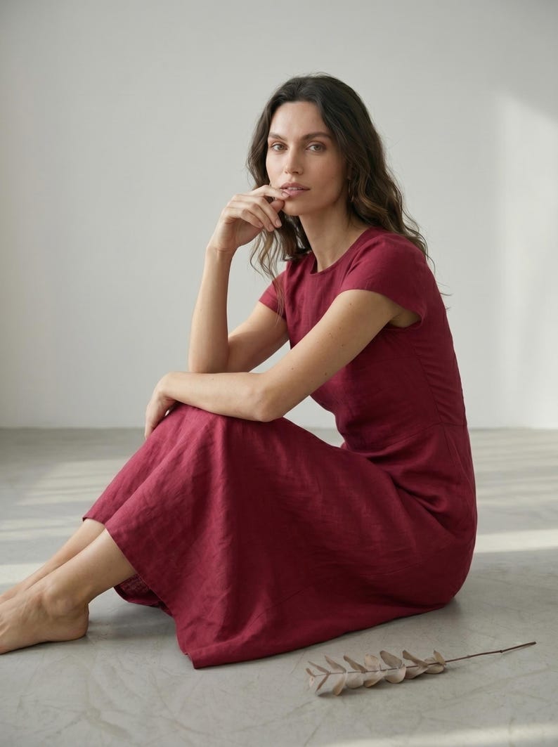 May include: A burgundy-colored dress with short sleeves. The dress has a round neckline and a flowing skirt. The model is sitting on a light gray surface. The dress appears to be made of linen or a similar fabric.