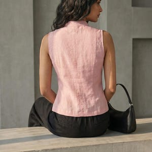 May include: A sleeveless, light pink linen top with a mandarin collar and vertical seams. The model is wearing black pants and a black shoulder bag. The top is a soft, muted pink color, and the overall style is minimalist and elegant.