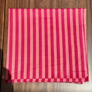 Op de afbeelding: Een gevouwen textiel met een verticaal streepjespatroon in roze tinten. De afwisselende strepen zijn een donkerdere en lichtere tint roze, wat een gedurfd visueel effect cre&euml;ert. De stof lijkt een lichtgewicht materiaal te zijn.