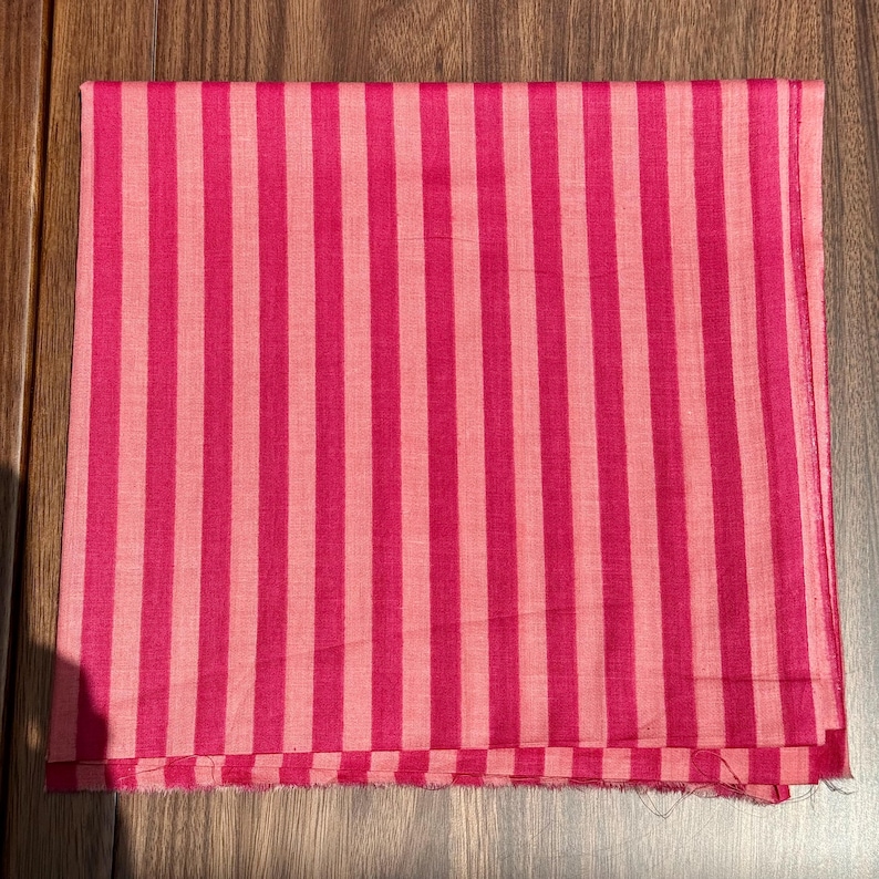 Op de afbeelding: Een gevouwen textiel met een verticaal streepjespatroon in roze tinten. De afwisselende strepen zijn een donkerdere en lichtere tint roze, wat een gedurfd visueel effect cre&euml;ert. De stof lijkt een lichtgewicht materiaal te zijn.