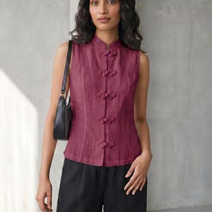 May include: A sleeveless, burgundy top with a mandarin collar and decorative frog closures. The top is paired with black pants and a black shoulder bag. The outfit is simple and elegant.
